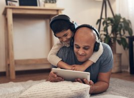 Een man en een jongen liggen op de grond en kijken samen naar een e-reader. De jongen heeft een koptelefoon op. Ze lachen.