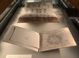 Een lange hal met in het midden glazen vitrines met opengeslagen boeken, aan de zijkanten rijen van bustes.