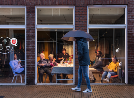 Mensen zitten in een warme kamer aan tafel, een voetganger loopt voorbij met paraplu in de hand.