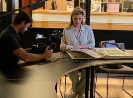 Presentatrice Welmoed Sijtsma bladert in een krant in de KB terwijl een cameraman haar filmt.