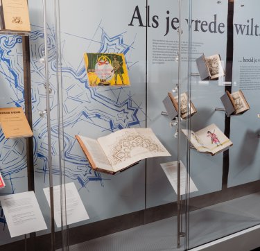 Een vitrine met boeken over oorlog en vrede en de tekst "Als je vrede wilt..." op de achtergrond.