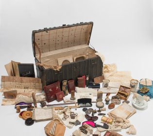 Miniatuurkist van de familie Cornets de Groot. De kist staat open en ervoor liggen spullen uitgestald, waaronder een landkaart en een aantal kleinere doosjes..