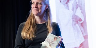 Conservator Karin Vingerhoets ontvangt een met kant ingepakt exemplaar van Zoeken naar Isah van Dido Michielsen. Fotograaf: Jacqueline van der Kort.