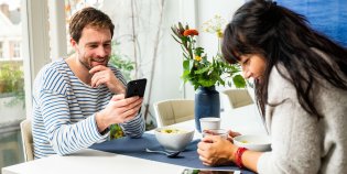 Man en vrouw lezend aan ontbijttafel. Man leest van telefoon. Vrouw leest van tablet.