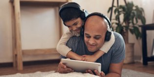 Een man en een jongen liggen op de grond en kijken samen naar een e-reader. De jongen heeft een koptelefoon op. Ze lachen.