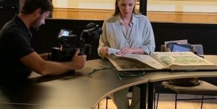 Presentatrice Welmoed Sijtsma bladert in een krant in de KB terwijl een cameraman haar filmt.