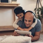 Een man en een jongen liggen op de grond en kijken samen naar een e-reader. De jongen heeft een koptelefoon op. Ze lachen.