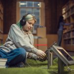 Jongere zit in een bibliotheek op de grond en kijkt op een tablet.