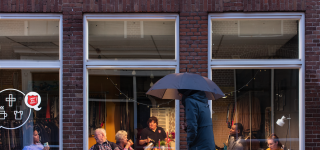 Mensen zitten in een warme kamer aan tafel, een voetganger loopt voorbij met paraplu in de hand.